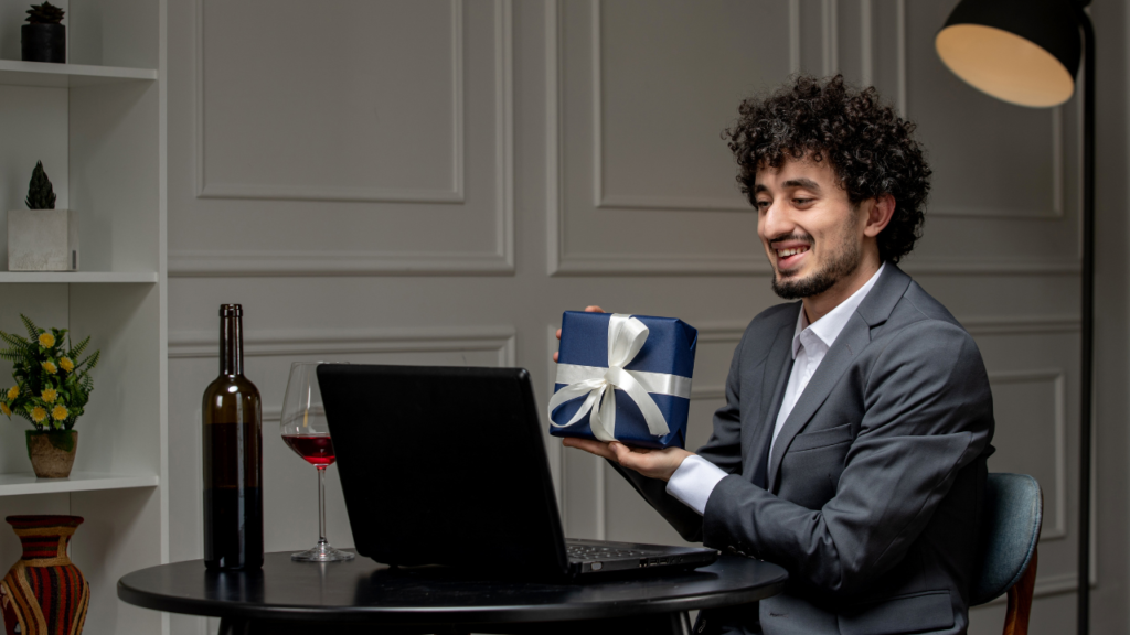 A man in a suit holds a gift box in one hand and a laptop in the other, smiling at the camera.