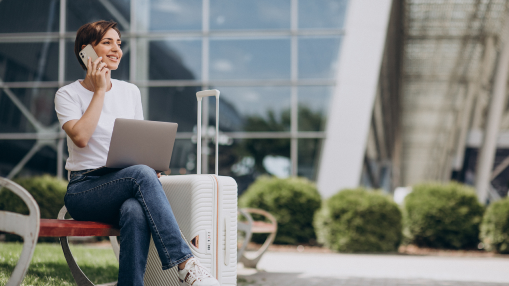 A woman sits on a bench with a laptop open beside a suitcase.