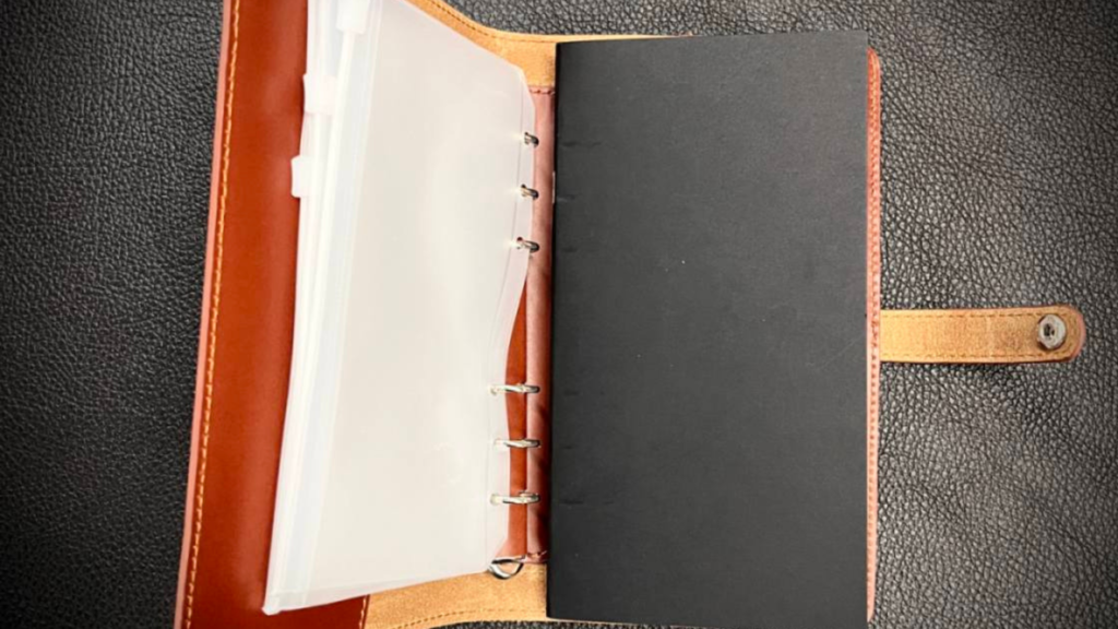 Open leather planner with a ring binder and empty plastic sleeves on the left, blank black divider on the right, laid on a textured gray surface.