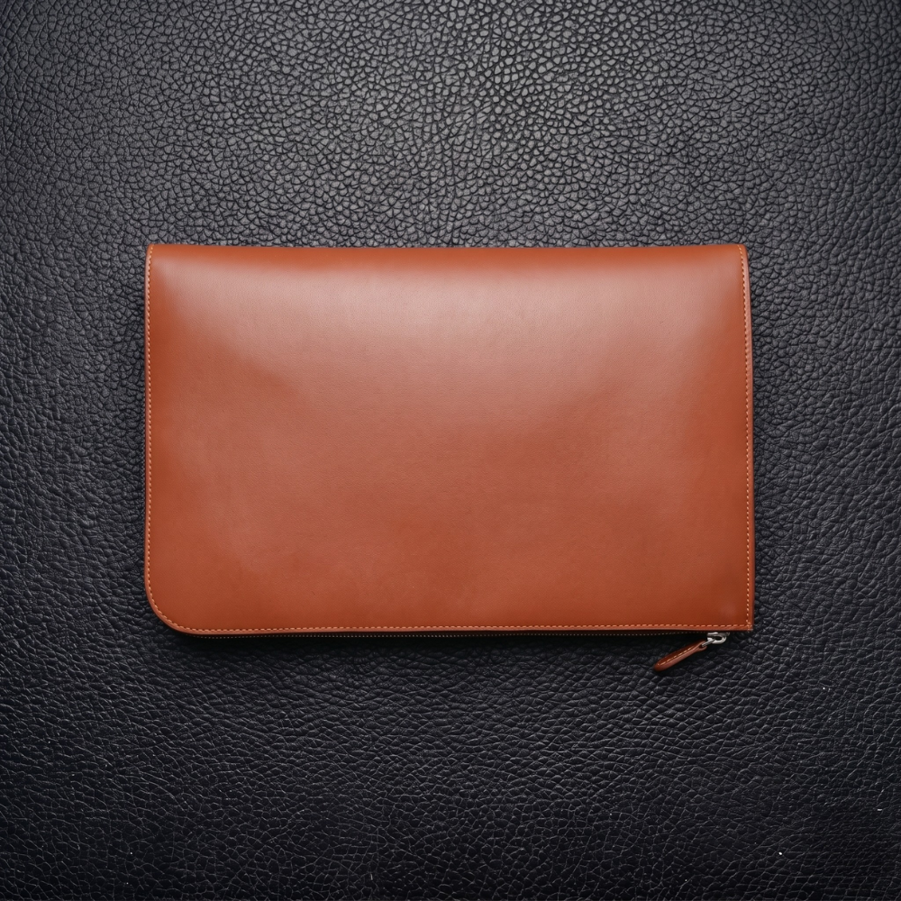 A sleek brown leather folio with a visible zipper on a textured black surface