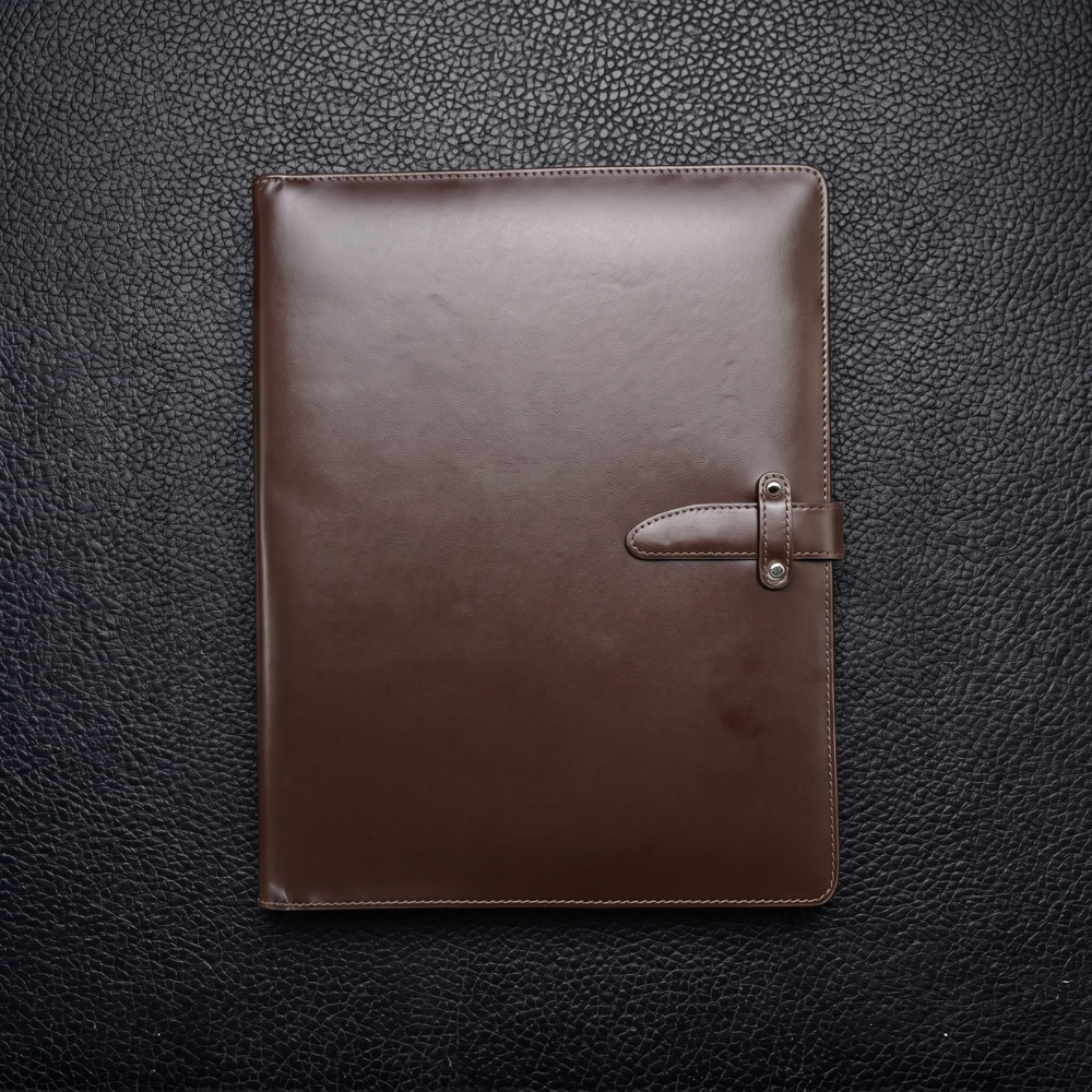 A sleek, closed brown leather meeting folder with a buckle, resting on a textured black leather surface.