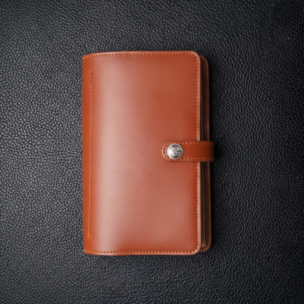 A sleek, brown leather notebook cover with a buttoned strap closure lies on a textured black surface