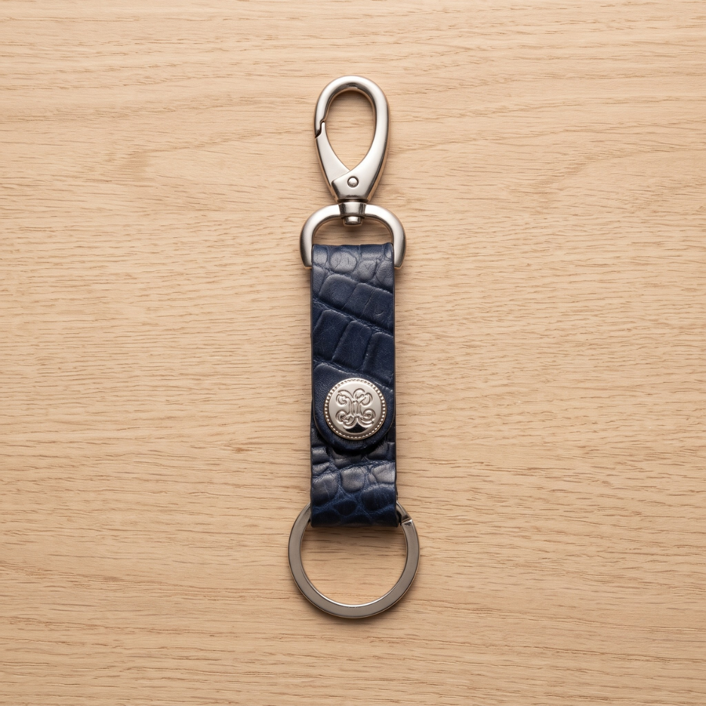 A luxury dark navy blue crocodile-patterned leather keychain strap fitted with a heavy-duty silver carabiner clip, a circular metallic logo rivet accent, and a flat split ring on a light wood backdrop.