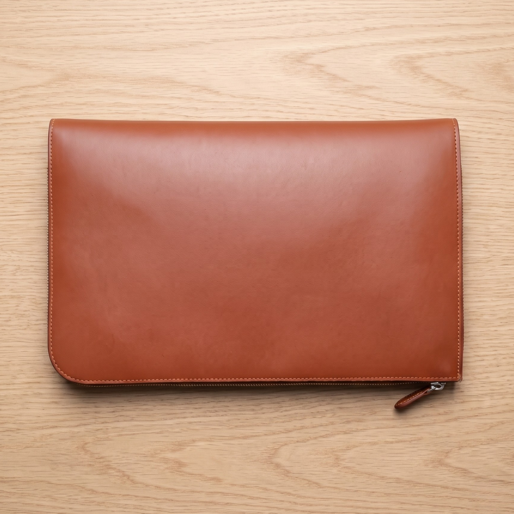 A flat-lay view of a minimalist, smooth bridle leather laptop folio with a secure perimeter zipper closure lying flat on a wooden grain tabletop.