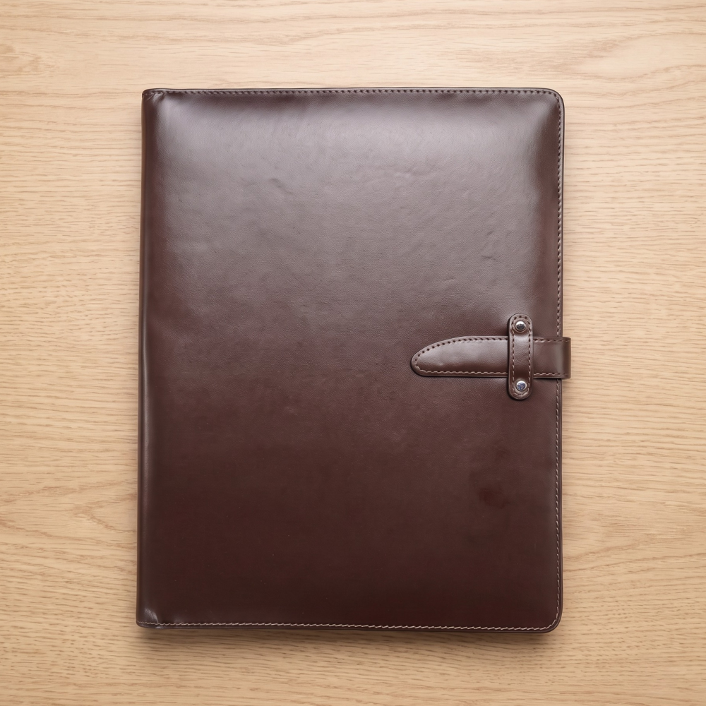 A premium dark brown smooth leather executive meeting folder with a built-in leather strap enclosure button closure system positioned vertically on a wood desk.