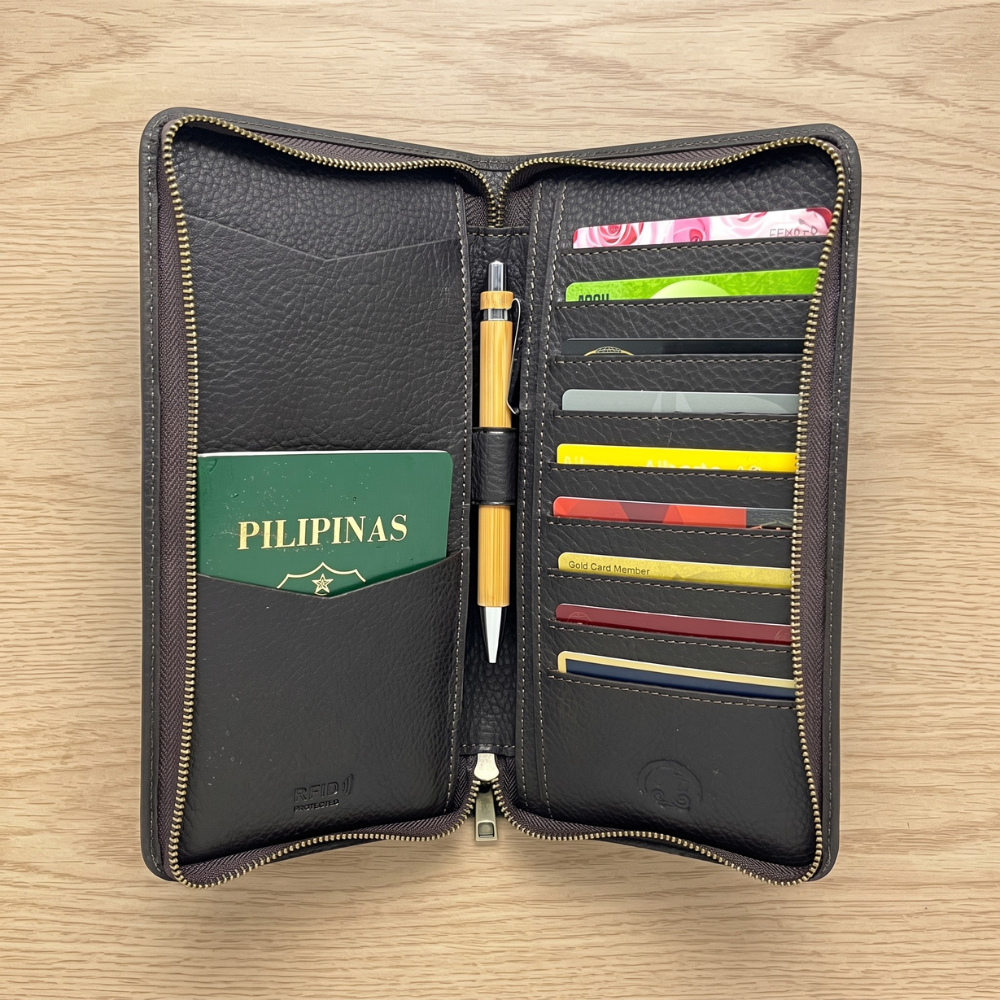 An open dark brown textured genuine leather travel wallet showing a green Philippine passport, slots filled with credit cards, an RFID protection stamp, and a centered wooden pen resting on a light wooden table background.
