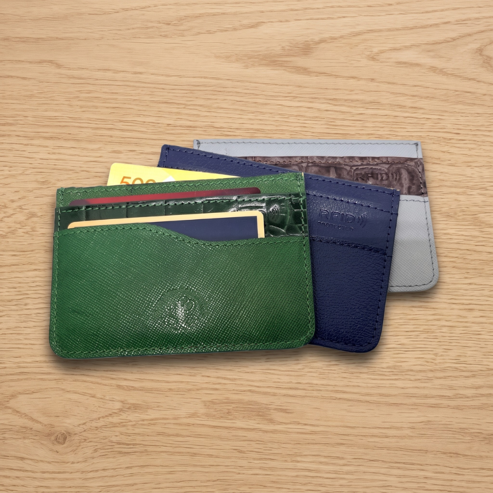 Three slim leather card holders with credit cards stacked diagonally on a wood desk, showcasing green saffiano, dark navy textured blue, and gray faux-crocodile leather patterns with embossed RFID protection icons.