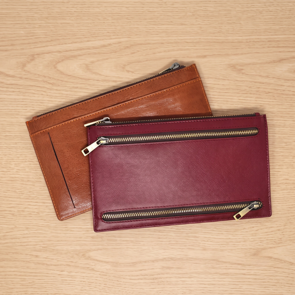 Two flat leather multi-currency cases with zipper closures stacked on a wooden surface.