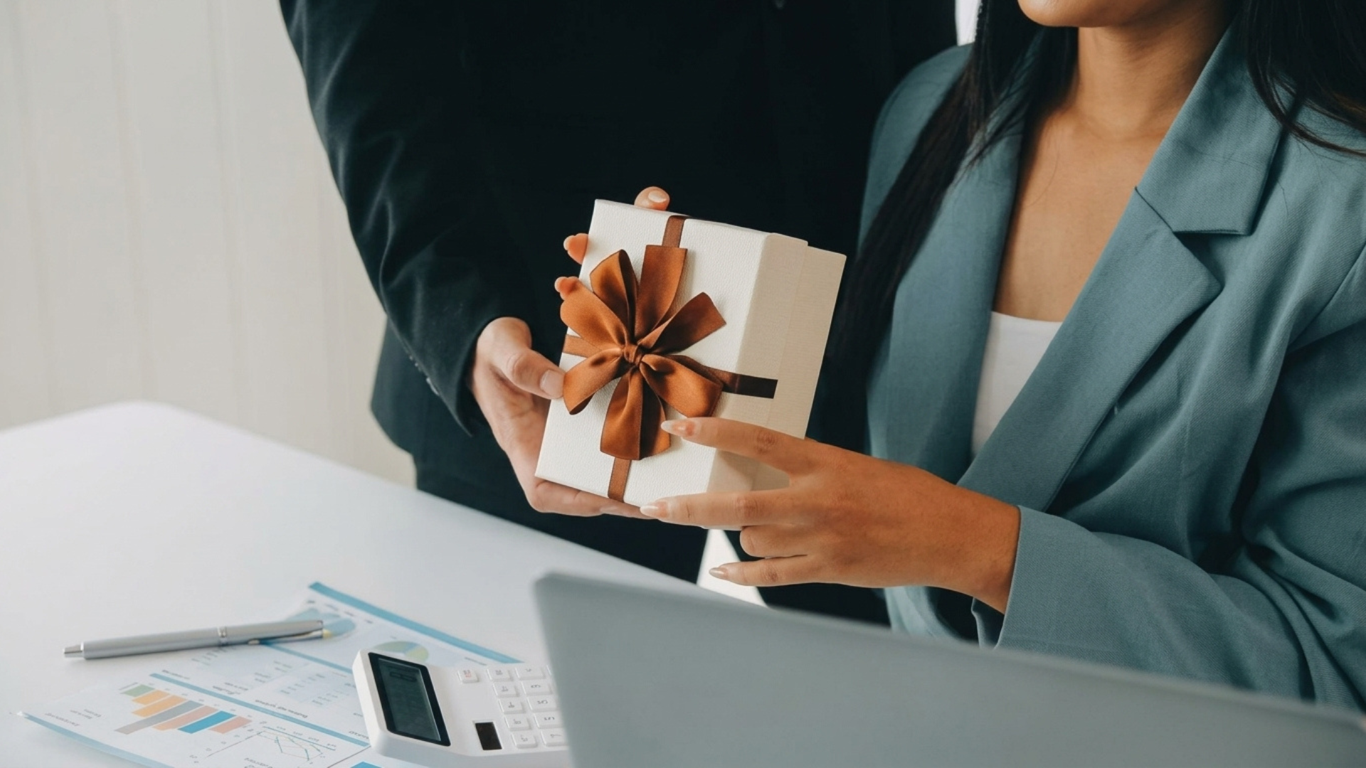 A businessman handing a white corporate gift box with a brown ribbon to a businesswoman across an office desk.